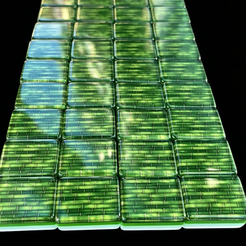 May include: A grid of square glass tiles arranged in a rectangular formation. Each tile features a vibrant green color with a horizontal bamboo-like pattern printed on the surface. The tiles have a glossy, reflective finish and smooth edges.