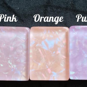 May include: Three rectangular cabochons with a smooth, glossy finish and an iridescent, marbled texture. The items are labeled with the text Pink, Orange, and Purple, corresponding to their respective light pink, peach, and light purple color tones.