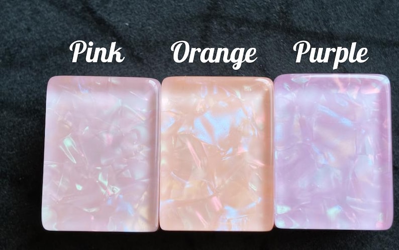 May include: Three rectangular cabochons with a smooth, glossy finish and an iridescent, marbled texture. The items are labeled with the text Pink, Orange, and Purple, corresponding to their respective light pink, peach, and light purple color tones.