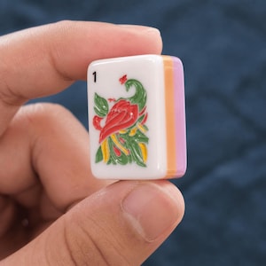 May include: A rectangular white mahjong tile featuring a colorful graphic of a bird with red, green, and yellow plumage. The top left corner displays the number 1 in black. The side of the tile shows stacked orange and light purple layers.