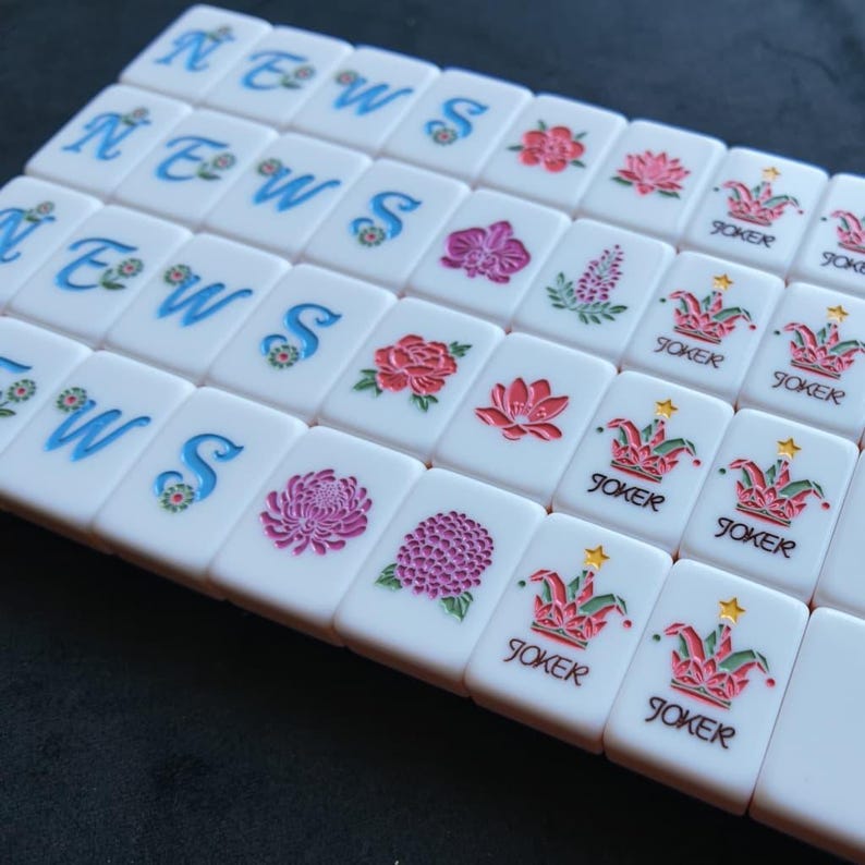 May include: A collection of white rectangular mahjong tiles arranged in rows. Some tiles feature the blue letters N, E, W, S with floral accents, while others display colorful botanical illustrations of flowers and joker icons with a yellow star.