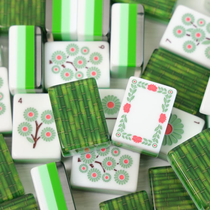 K&ouml;nnte beinhalten: Eine Sammlung von Mahjong-Spielsteinen, bestehend aus wei&szlig;en Steinen mit gr&uuml;n-roten Blumenmustern, gr&uuml;nen Bambusmustern und Steinen mit vertikalen gr&uuml;nen Streifen. Einige Steine zeigen die Zahlen 1, 4, 6 und 7 in schwarzer Schrift.