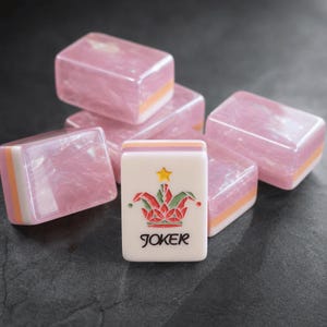 May include: A collection of rectangular mahjong tiles featuring a translucent pink marbled finish with white and orange layered bases. One tile stands upright in the foreground, displaying a colorful jester hat graphic, a yellow star, and the word JOKER.