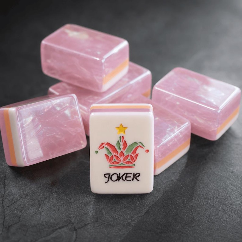 May include: A collection of rectangular mahjong tiles featuring a translucent pink marbled finish with white and orange layered bases. One tile stands upright in the foreground, displaying a colorful jester hat graphic, a yellow star, and the word JOKER.