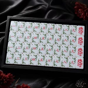 May include: A set of white rectangular Mahjong tiles arranged in a black tray. Each tile features red Chinese numerals from one to nine, accompanied by green floral vine illustrations. The final column displays tiles with a red dragon motif.