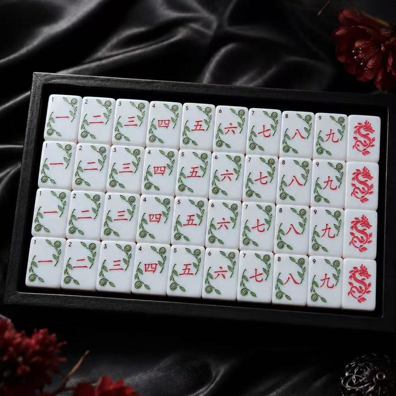 May include: A set of white rectangular Mahjong tiles arranged in a black tray. Each tile features red Chinese numerals from one to nine, accompanied by green floral vine illustrations. The final column displays tiles with a red dragon motif.