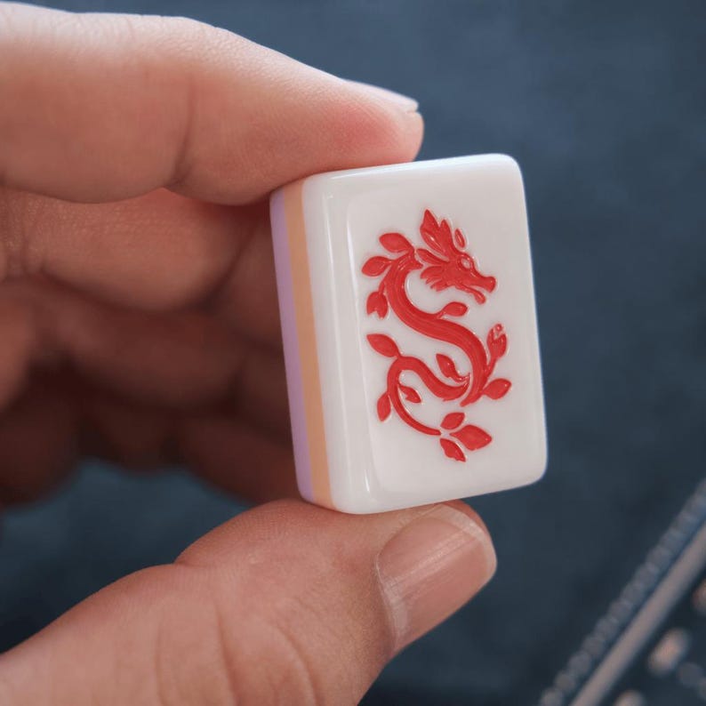 May include: A rectangular white Mahjong tile featuring a red engraved dragon design on its front face. The tile has a smooth, glossy finish with a thick profile, showing a layered construction with a light-colored side panel visible on the left edge.