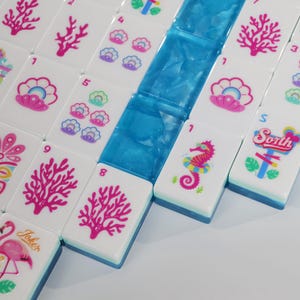 May include: A set of white Mahjong tiles featuring colorful ocean-themed graphics including pink coral, seashells, seahorses, and a flamingo. Some tiles display numbers and the text 'Sorth' or 'Joker'. A row of translucent blue tiles is included.