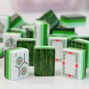 K&ouml;nnte beinhalten: Eine Sammlung rechteckiger Mahjong-Spielsteine in Gr&uuml;n und Wei&szlig;. Die Steine zeigen verschiedene Motive, darunter ein Bambusmuster, eine florale Umrandung sowie einen Spielstein mit der Ziffer zwei und zwei gr&uuml;nen Blumen-Symbolen.