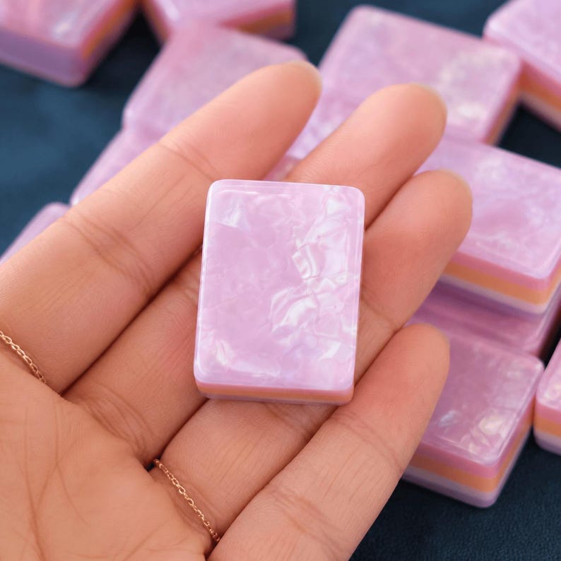May include: A rectangular mahjong tile featuring a glossy, marbled light pink top surface with a layered base consisting of a thin orange stripe sandwiched between two light purple sections, showcasing a smooth, polished finish throughout the piece.