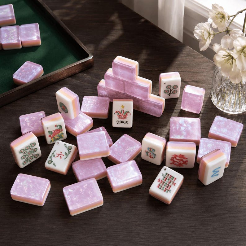 May include: A collection of rectangular mahjong tiles featuring a pearlescent lavender and white marbled finish. The tiles display various traditional symbols, numbers, and a central joker tile with a colorful crown graphic and the text JOKER.