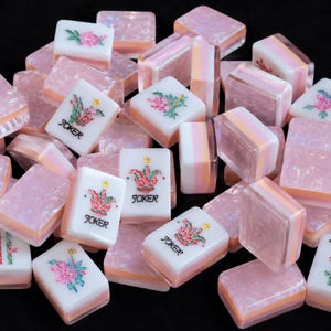 May include: A collection of rectangular mahjong tiles featuring a white face with colorful floral or Joker designs and text, paired with a translucent, pearlescent pink and peach layered base. The tiles are scattered in a pile, showing various orientations.