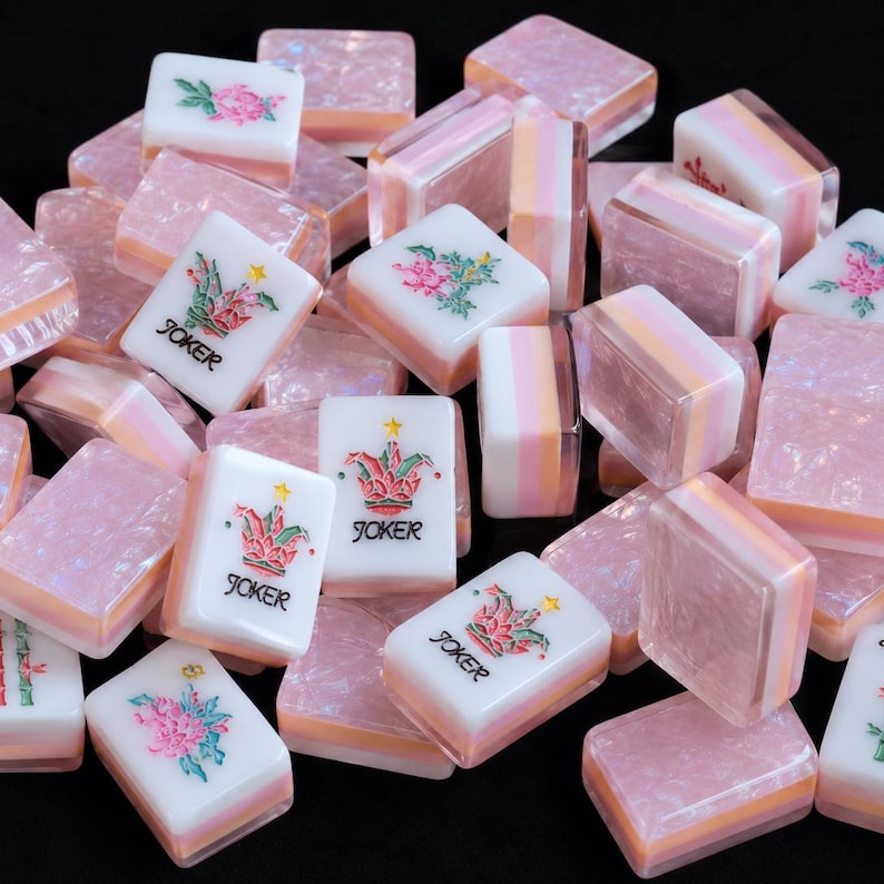 May include: A collection of rectangular mahjong tiles featuring a white face with colorful floral or Joker designs and text, paired with a translucent, pearlescent pink and peach layered base. The tiles are scattered in a pile, showing various orientations.