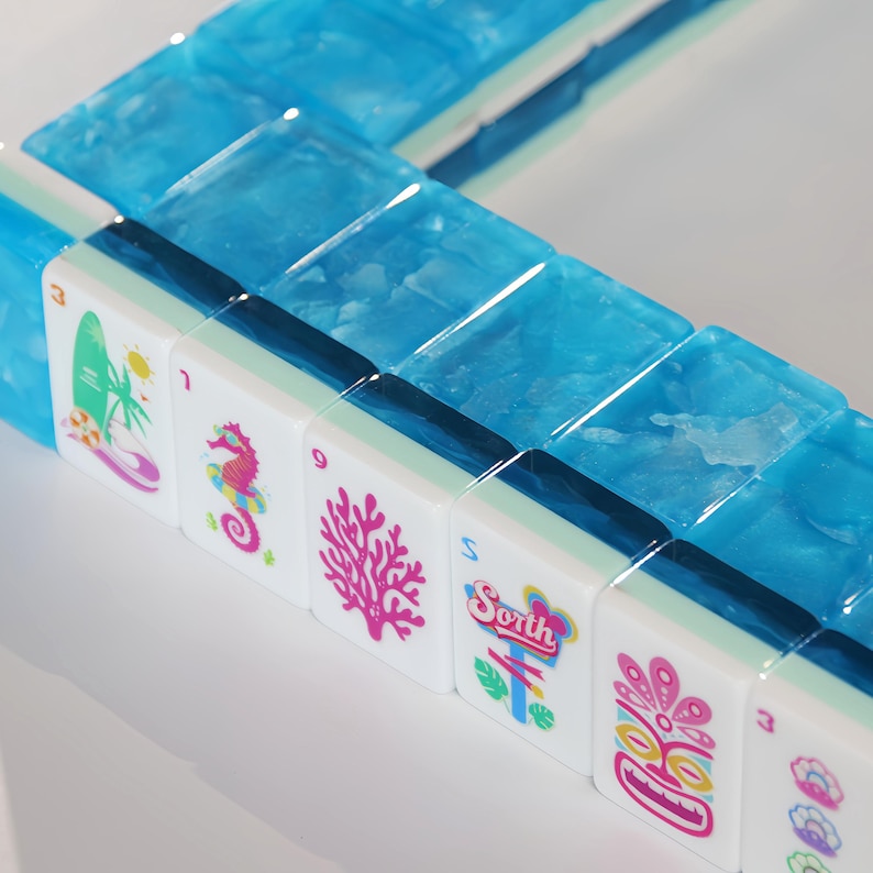 May include: A set of mahjong tiles featuring translucent blue backs with a textured finish. The white faces display colorful graphics including a surfboard, a seahorse, a pink coral, a sign reading South, a tiki mask, and a pearl in a shell.