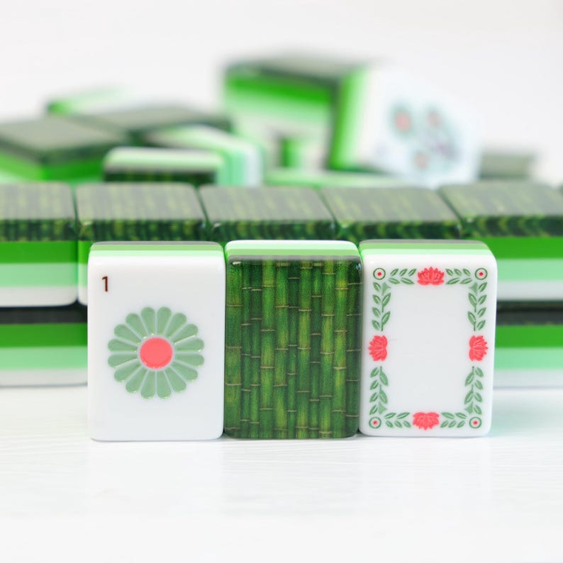 K&ouml;nnte beinhalten: Ein Set aus rechteckigen Mahjong-Spielsteinen in Gr&uuml;n und Wei&szlig;. Drei Steine befinden sich im Vordergrund: einer mit einer hellgr&uuml;nen Blume und der Zahl 1, einer mit einem gr&uuml;nen Bambusmuster und einer mit einem floralen Rahmen.