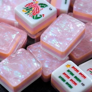 May include: A collection of mahjong tiles featuring a mix of iridescent pink cracked-texture surfaces and white tiles with colorful floral and bamboo engravings. The tiles have layered pink, orange, and white bases, with some displaying a small yellow star.