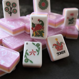 May include: A collection of rectangular Mahjong tiles featuring white faces with colorful engraved designs, including a peacock, a joker with a star, and floral motifs. The tiles have a layered base with a pink pearlescent top and orange middle.