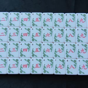 May include: A collection of white rectangular mahjong tiles arranged in a grid. Each tile features a red Chinese numeral from two to nine, accompanied by a green floral vine design. Small black Arabic numerals are printed in the top left corner.