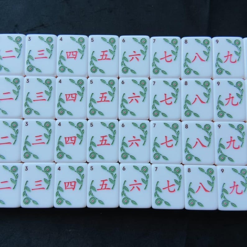 May include: A collection of white rectangular mahjong tiles arranged in a grid. Each tile features a red Chinese numeral from two to nine, accompanied by a green floral vine design. Small black Arabic numerals are printed in the top left corner.