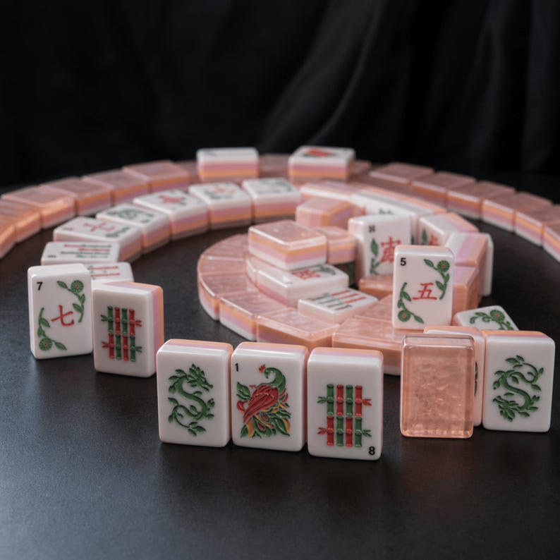 May include: A set of rectangular mahjong tiles arranged in a spiral pattern. The tiles are white with translucent peach-colored backs and feature engraved green, red, and black symbols, including Chinese characters, bamboo, dragons, and a bird.