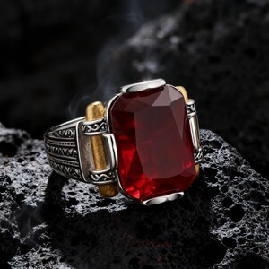 May include: A silver ring featuring a large, rectangular, deep red faceted gemstone set in the center. The band displays intricate engraved patterns on the sides, accented by two vertical gold-toned bars flanking the central red stone setting.