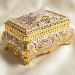 May include: Ornate jewelry box with a gold finish and intricate floral detailing. The box features a light purple base color with gold accents and a hinged lid. The box is rectangular with curved sides and small feet.