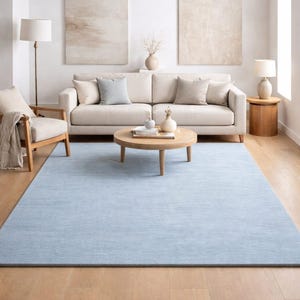 Könnte beinhalten: Hellblauer rechteckiger Teppich in einem Wohnzimmer. Der Teppich liegt auf einem Holzboden, mit einem beigen Sofa, einem Holztisch und zwei abstrakten Gemälden an der weißen Wand. Ein Holzstuhl und eine Lampe sind ebenfalls zu sehen.
