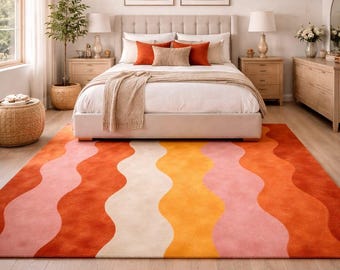 May include: A bedroom featuring a large, colorful rug with a wavy pattern in shades of orange, pink, and cream. The rug sits on a light wood floor, complementing the neutral tones of the room's furniture and decor. The room includes a bed, nightstands, and artwork.