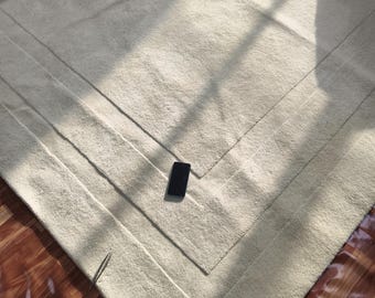 May include: A large, off-white area rug with a textured, tufted surface and a subtle, recessed rectangular border design. A small, black rectangular smartphone is placed centrally on the rug, providing a sense of scale to the floor covering.