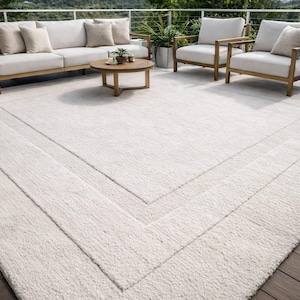 Op de afbeelding: Een groot, crèmekleurig buitenkleed met een getextureerd oppervlak en een geometrische rand. Het kleed ligt op een houten terras en is omgeven door tuinmeubelen, waaronder een bank, stoelen en een salontafel. Het kleed meet ongeveer 244 x 305 cm.