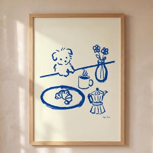 Puede incluir: Una lámina enmarcada con un dibujo de líneas azules estilo tinta sobre fondo crema. La ilustración muestra a un perro pequeño sentado a una mesa con un cruasán en un plato, una taza humeante, una cafetera italiana y un jarrón con dos flores.
