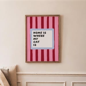 May include: A framed print with a pink and red striped background. The print features a light blue rectangle with the words "HOME IS WHERE MY CAT IS" in black text. The frame is a natural wood color.