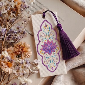 May include: A rectangular bookmark made of off-white fabric featuring a cross-stitched design of a central pink and purple lotus flower surrounded by intricate teal and yellow scrollwork, finished with a dark purple silk tassel at the top.