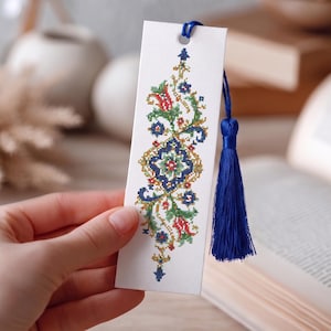 May include: A rectangular white bookmark featuring a vertical cross-stitch pattern in shades of blue, green, red, and yellow. The design includes floral and geometric motifs. A vibrant blue silk tassel is attached to the top through a punched hole.