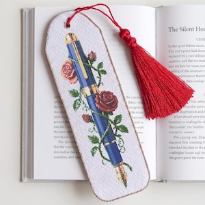 Peut inclure: Un marque-page en tissu blanc orné d'un motif au point de croix représentant un stylo plume bleu entrelacé de roses rouges et de feuilles vertes. Il présente un bord cousu beige, un sommet arrondi et un long pompon décoratif rouge vif.