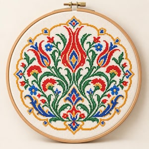 May include: A circular wooden embroidery hoop containing a cross-stitch pattern on white fabric. The design features a symmetrical floral motif with red, green, blue, and yellow threads, arranged in a central tulip shape surrounded by leaves.