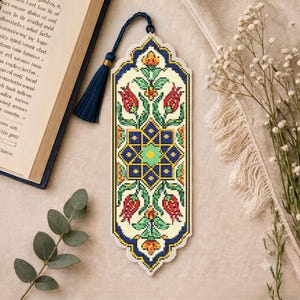 May include: A rectangular bookmark featuring a cross-stitch pattern with a central blue and yellow geometric star motif, surrounded by red tulips and green foliage on a cream background, finished with a dark blue tassel at the top.