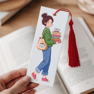 May include: A white bookmark featuring an illustration of a person carrying books and a tote bag with a coffee cup design. The bookmark has a red tassel and a small hole at the top. The person is wearing a green sweater, blue jeans, and pink shoes.