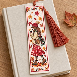 May include: A rectangular cross-stitch bookmark on a light fabric background featuring a person in a red hat, jumper, and skirt holding a mug, surrounded by falling autumn leaves in shades of red, orange, and yellow, finished with a rust-coloured tassel.