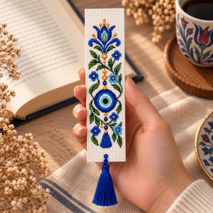 May include: A rectangular white bookmark featuring a cross-stitch pattern of a blue evil eye symbol, green leaves, and blue flowers. The design includes a vertical arrangement of floral motifs and a dangling blue tassel attached to the bottom edge.