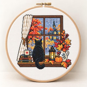 Puede incluir: Un bastidor de bordado circular de madera con un patrón de punto de cruz de un gato negro sentado en un alféizar. La escena muestra una vista otoñal lluviosa, libros, una taza con un corazón, una linterna encendida y una calabaza.