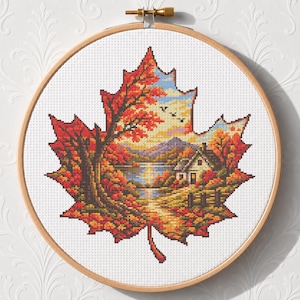 May include: A circular wooden embroidery hoop containing a cross-stitch pattern shaped like a large maple leaf. The design features a landscape with a small house, a lake, mountains, and trees with vibrant red, orange, and yellow autumn foliage.