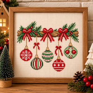 May include: A framed cross-stitch embroidery on cream fabric featuring five hanging Christmas ornaments in red and green. The ornaments are suspended from a horizontal wooden branch decorated with green pine needles, red berries, and three red bows.