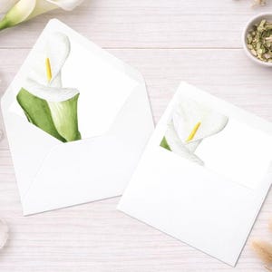May include: Two white envelopes featuring watercolour calla lily illustrations. The flowers have white petals, yellow stamens, and green leaves. The envelopes rest on a light wood-grain surface, suitable for stationery or invitations.