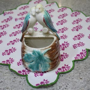 May include: A ceramic wall pocket featuring two birds perched together. The birds have white bodies, teal wings, and yellow heads. The pocket is brown and cream-colored with a teal leaf design. The pocket is sitting on a white floral patterned placemat.