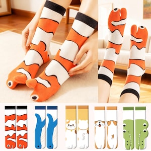 May include: Orange, white, and black novelty socks with a clownfish design. The socks have a split toe and feature a cartoon-like fish face on the foot. Additional pairs of socks with animal designs are also shown.