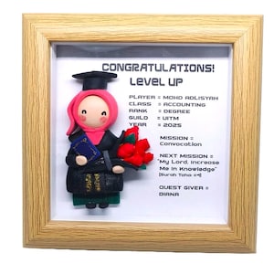 Op de afbeelding: Een ingelijst afstudeercadeau met een kleifiguur van een afgestudeerde in een zwarte toga en hoed, met een boeket en een diploma. De lijst bevat de tekst "CONGRATULATIONS! LEVEL UP" en details over de graad en toekomstige doelen van de afgestudeerde.