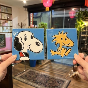 May include: Two square ceramic tiles with cartoon character designs. One tile features Snoopy with a black ear and red collar on a blue background. The other tile has a yellow Woodstock character on a blue background.