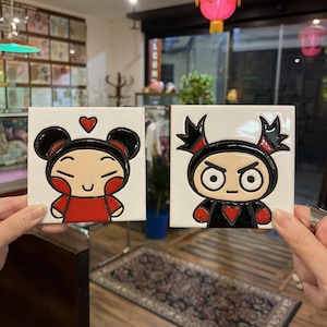 May include: Two square ceramic tiles with cartoon characters. One tile shows a smiling character in red and black, with a heart above. The other tile depicts an angry character, also in red and black.