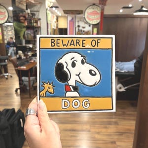 May include: Square ceramic tile featuring Snoopy from Peanuts with the text "BEWARE OF DOG". The tile has a blue background with a yellow border. Snoopy is white with black ears and a red and black collar. A small yellow Woodstock is also present.